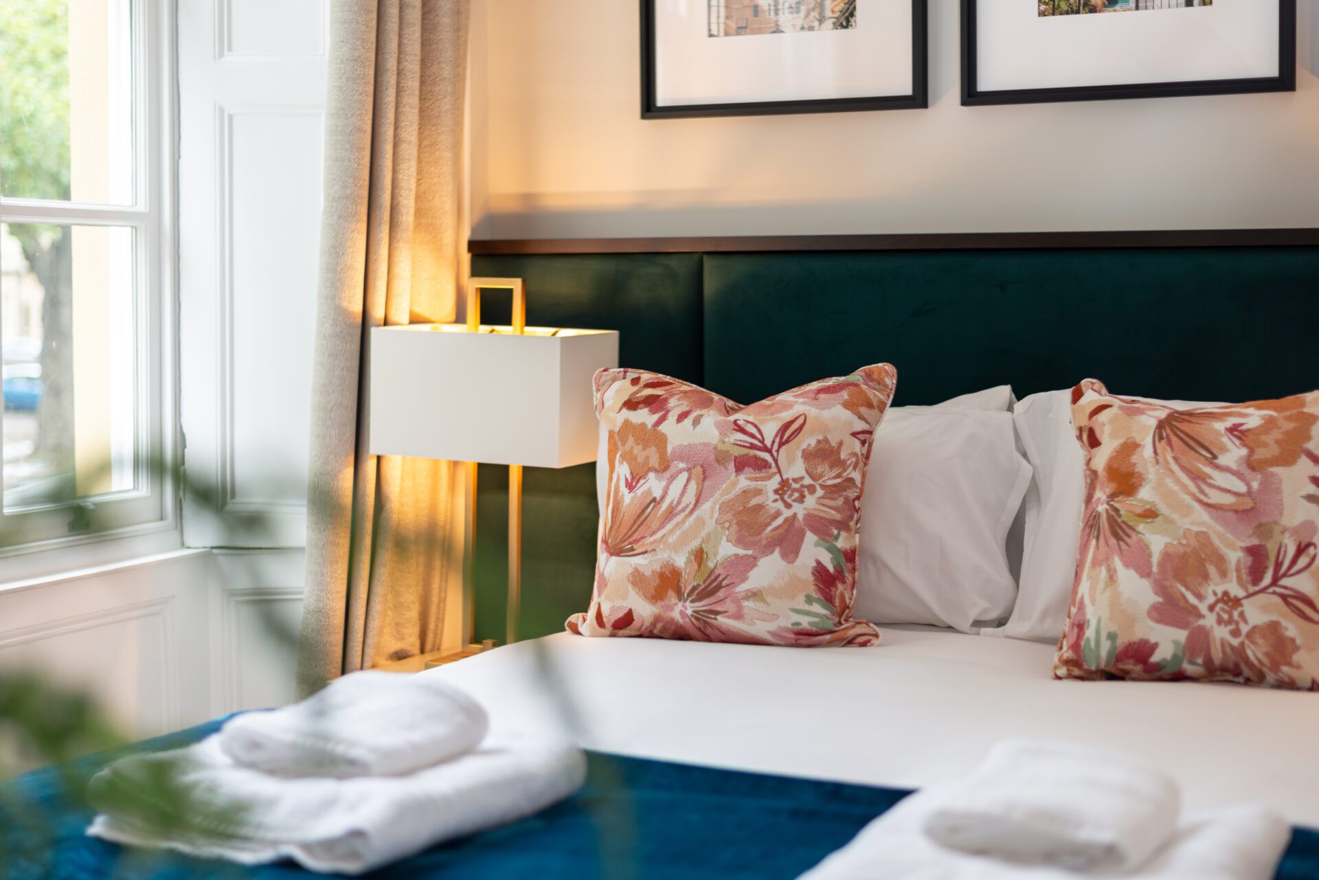 Close up of well presented bed with red patterned cushions with navy blue headboard. Bedside lamp and table to the left by a sash window.
