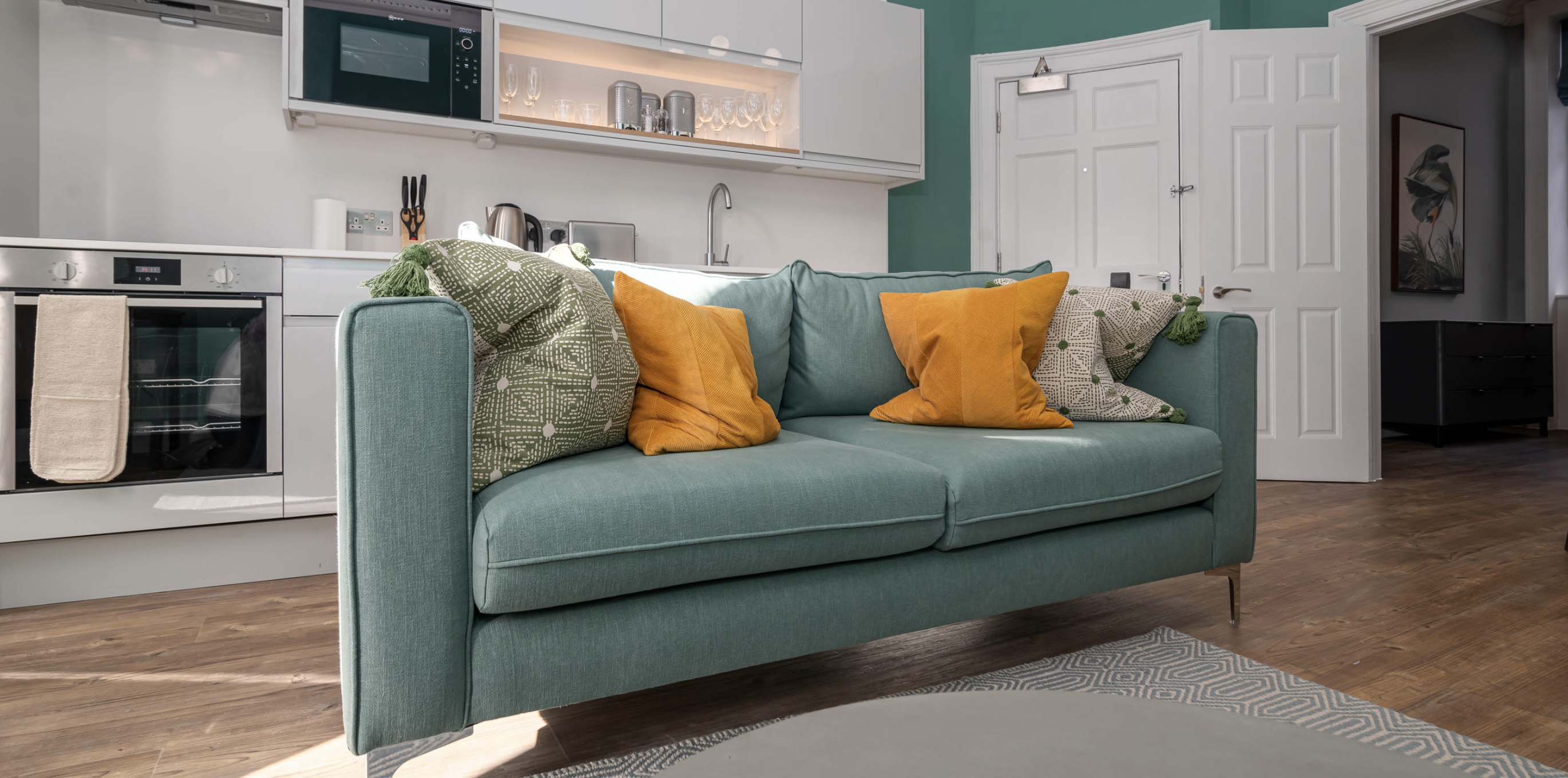 close up of green sofa with mustard yellow cushions and kitchenette behind