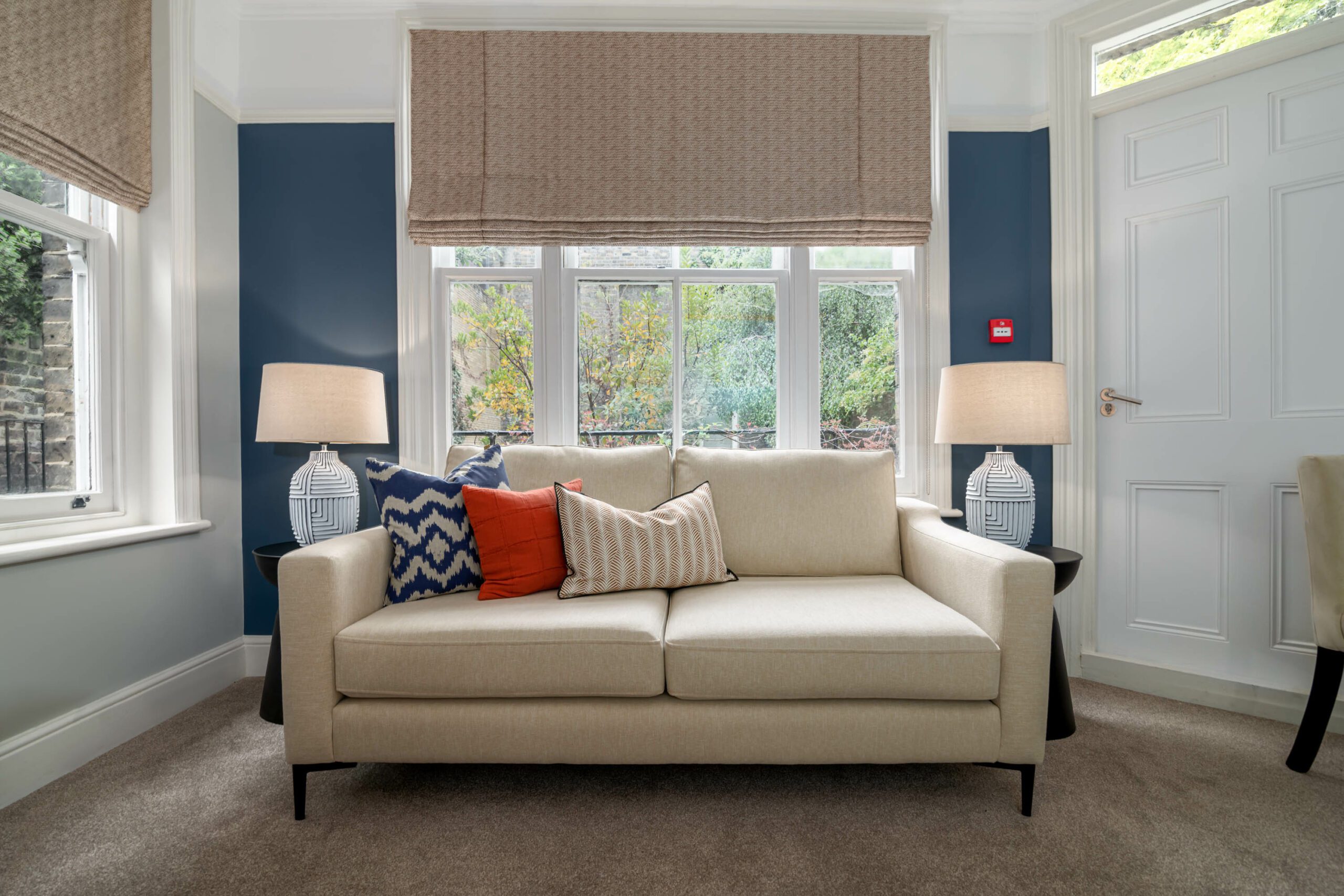 Cream sofa in front of window with red and blue cushions. Blue feature wall behind and 2 side lamps