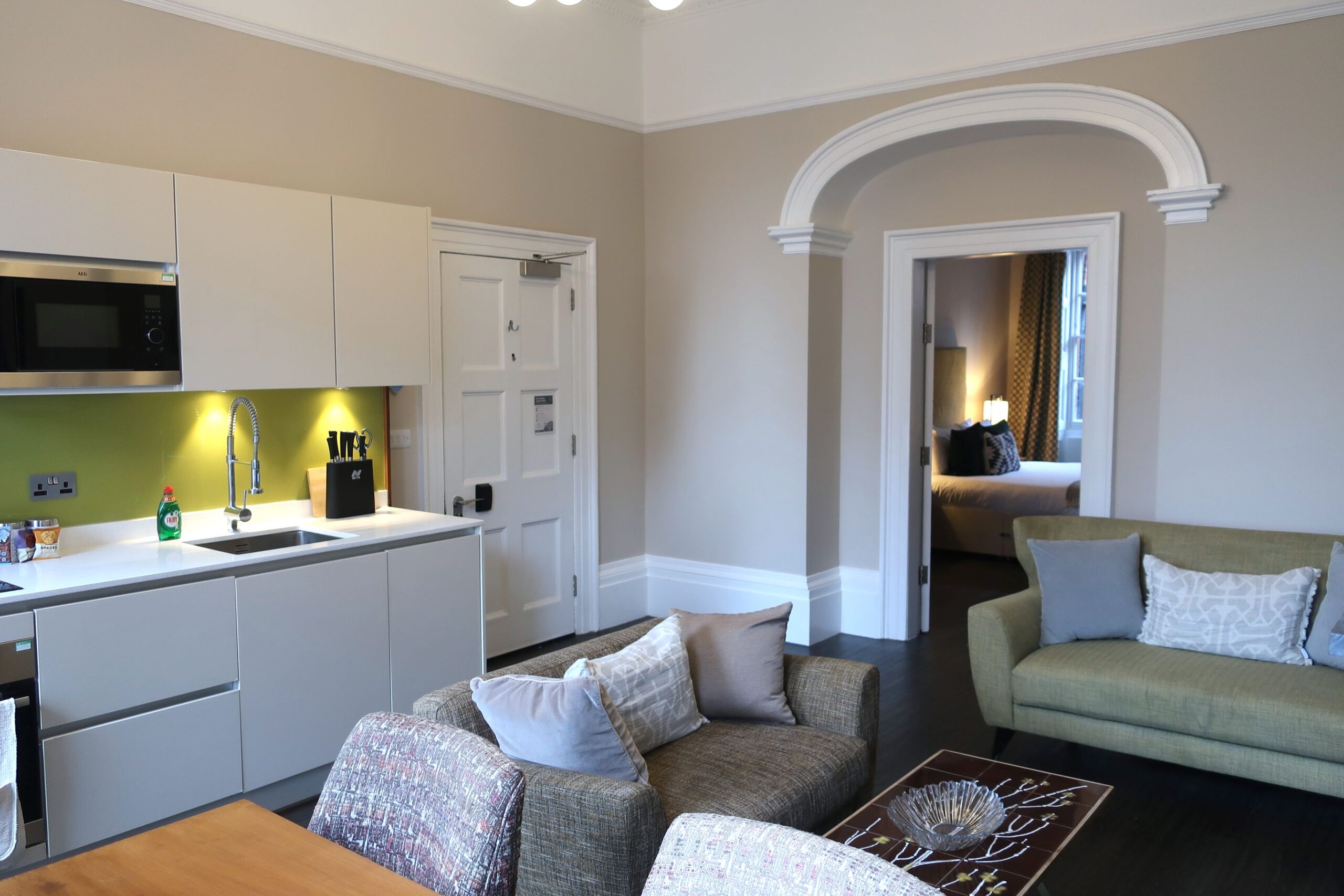 A living room displaying a couch, a table, a microwave, and an open kitchen area in the background.